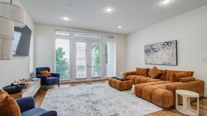 A spacious living room with a large sectional sofa, a blue armchair, and a coffee table. There is a large window with blinds, a painting on the wall, and a ceiling light fixture. - Hermann Park Hideaway Sleeps 12 w/ Parking & Patio (Houston)