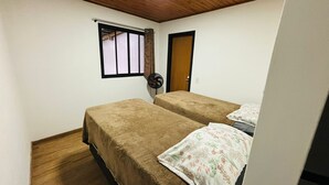 A small, single-room accommodation with two beds, a window, a door, and a ceiling fan.