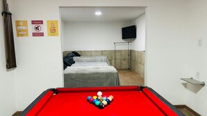 A pool table with balls in the foreground, a bed with a grey blanket, and a TV mounted on the wall.