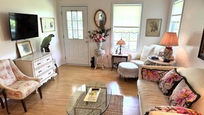 A living room with a glass coffee table, floral armchairs, a sofa with floral cushions, a white dresser, a television, a door with a stained glass window, and a vase with flowers. - Charming 2BR Cottage on The Point (Colonial Beach)