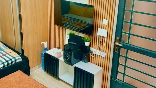 A compact room with a bed, a TV mounted on the wall, a small table with a speaker, and a radiator. - G1 Studio with a lift,balcony &underground parking (Nairobi)