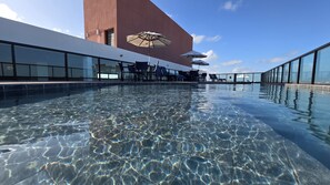 A clear indoor swimming pool with lounge chairs and umbrellas, surrounded by glass walls and a modern building. - Flat Porto de galinha Villas 311 byLP (Ipojuca)