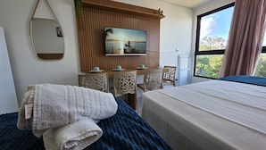 Habitación de hotel con cama, escritorio con sillas, televisor montado en un panel de madera y ventana con vista a follaje.