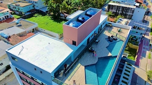 Un hotel de varios pisos con piscina en la azotea, un área exterior techada y un edificio grande con tanques de agua azules.