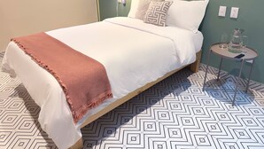 A neatly made bed with a white and pink blanket, a bedside lamp, a small table with a glass pitcher and glasses, and a geometric patterned floor. - Private room with private bathroom in the Americana neighborhood (Guadalajara)