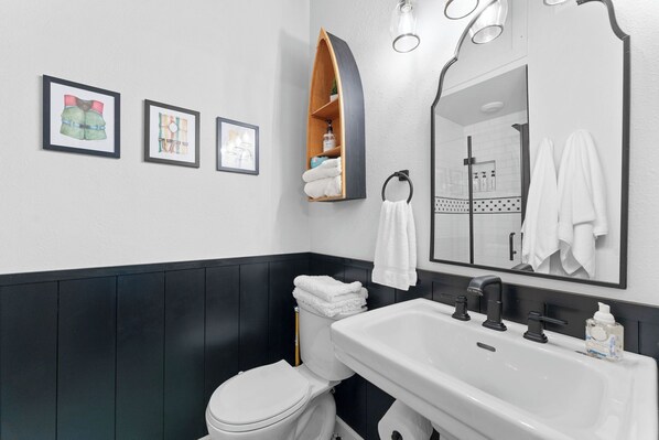 Un baño moderno con un lavamanos blanco, grifos negros, un inodoro y un espejo con un sujetapapeles. Hay tres cuadros enmarcados en la pared.