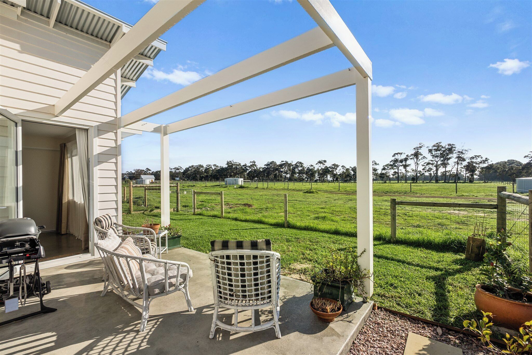 A covered patio with outdoor furniture, a grill, and a view of a grassy field and trees.