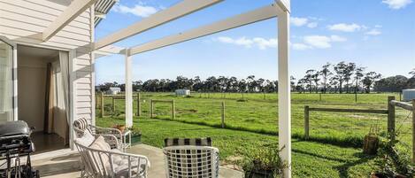 A covered patio with outdoor furniture, a grill, and a view of a grassy field and trees.
