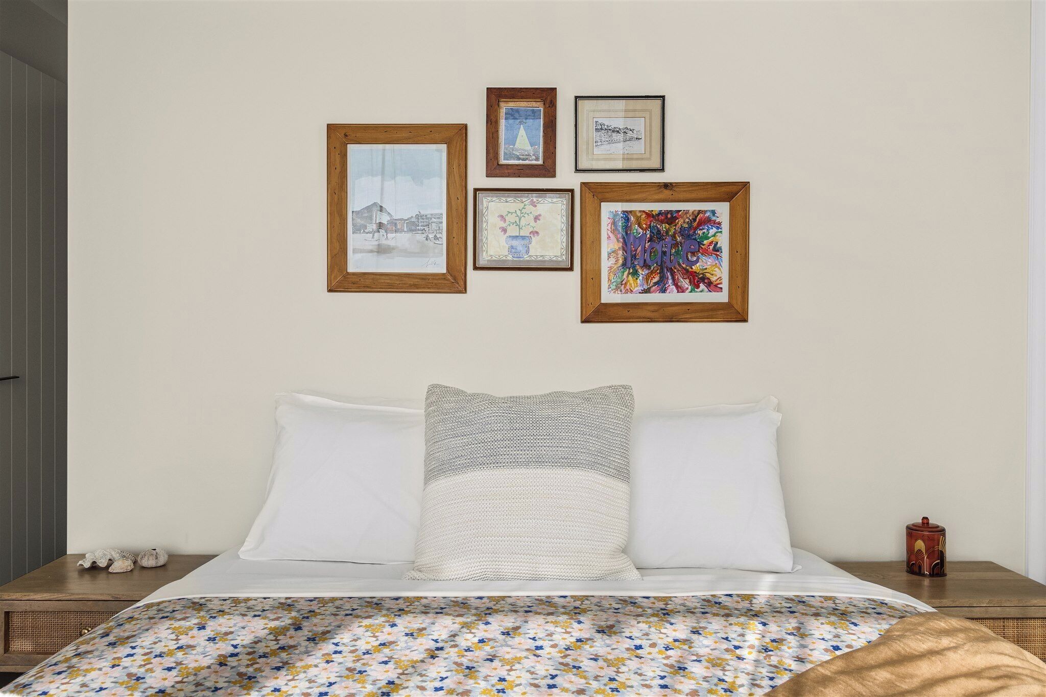 A neatly made bed with a floral-patterned bedspread, a grey textured pillow, and a white pillow. There are four framed artworks on the wall above the bed.