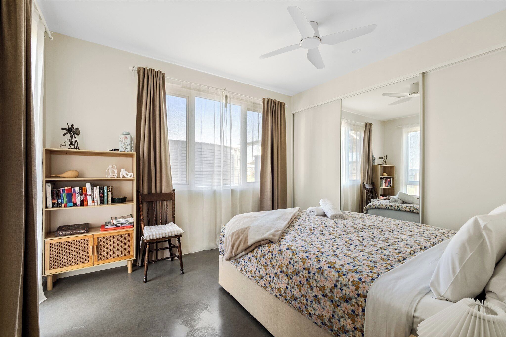 A bedroom with a bed, a bookshelf, a chair, a ceiling fan, and a large mirror.