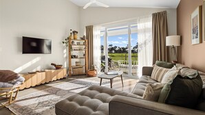Ein Wohnzimmer mit einer Couch, einem Sessel und einem Fernseher an der Wand. Ein großes Fenster mit Vorhängen bietet Blick auf viel Grün draußen.