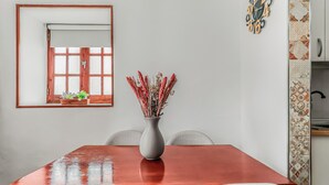 Ein Essbereich mit Holztisch, weißen Stühlen, einer Vase mit Trockenblumen, einem Fenster mit Aussicht und einer Wanduhr.