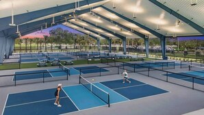 Un court de tennis intérieur comprenant plusieurs terrains, un espace de repos couvert et une vue sur le parc environnant.
