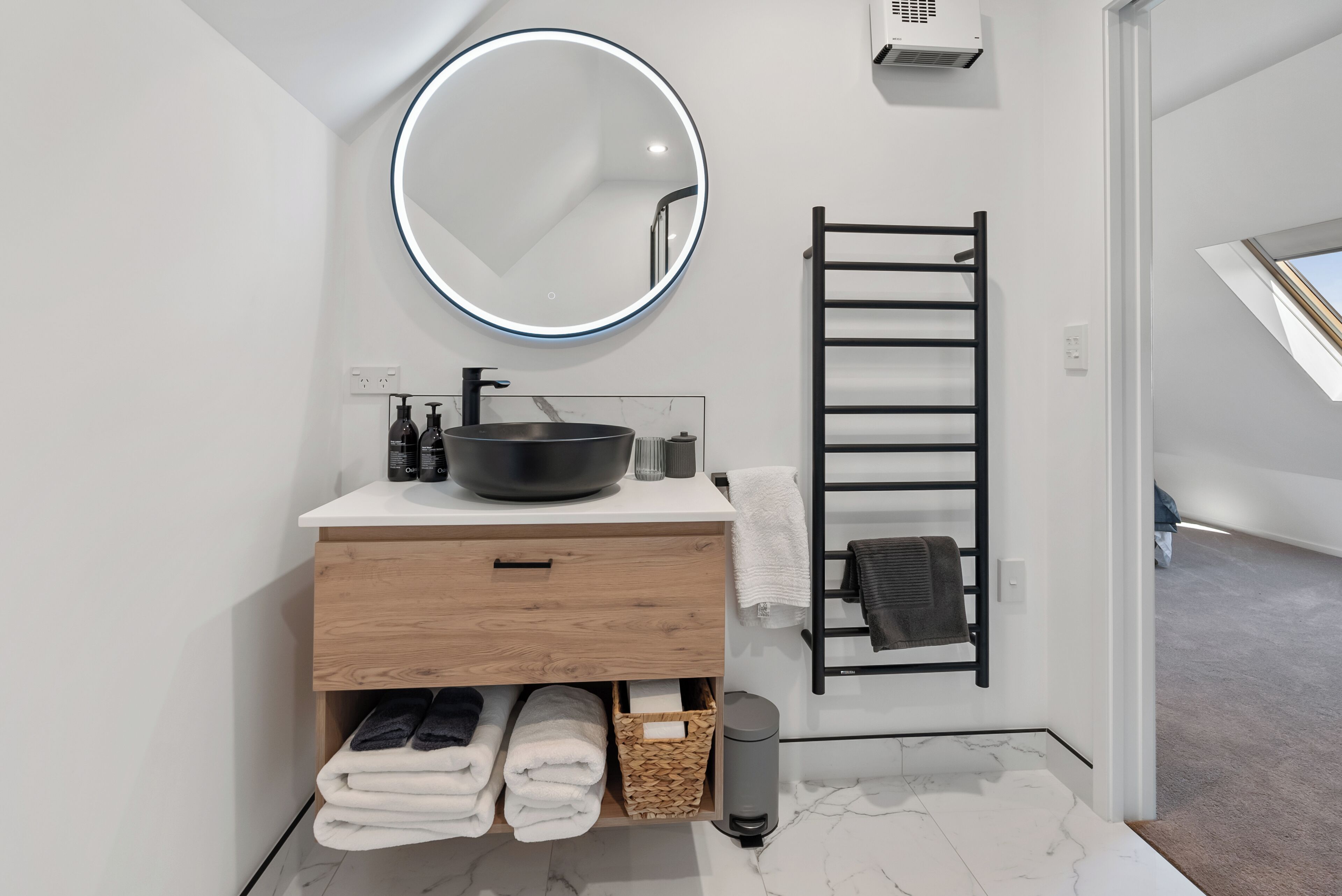 Een moderne badkamer met een ronde spiegel, een zwarte wastafel, een handdoekhouder en een houten opbergmeubel.