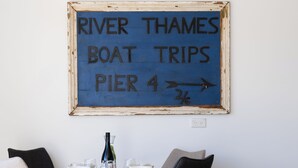 A room with a table set for two, a chair, a sofa, and a sign indicating a boat trip to River Thames Pier 4.