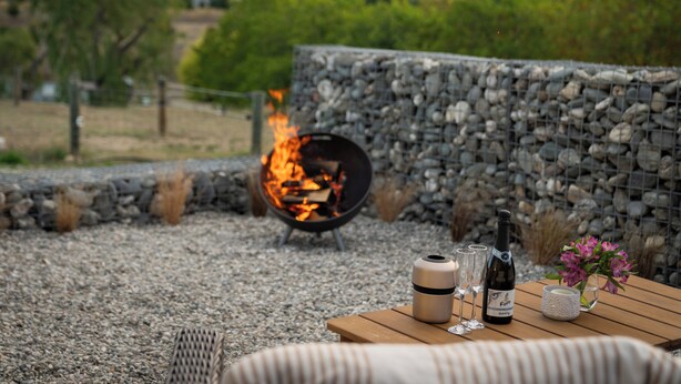 A cozy outdoor seating area with a fire pit, a table with a bottle and glasses, and a view of the mountains in the background.