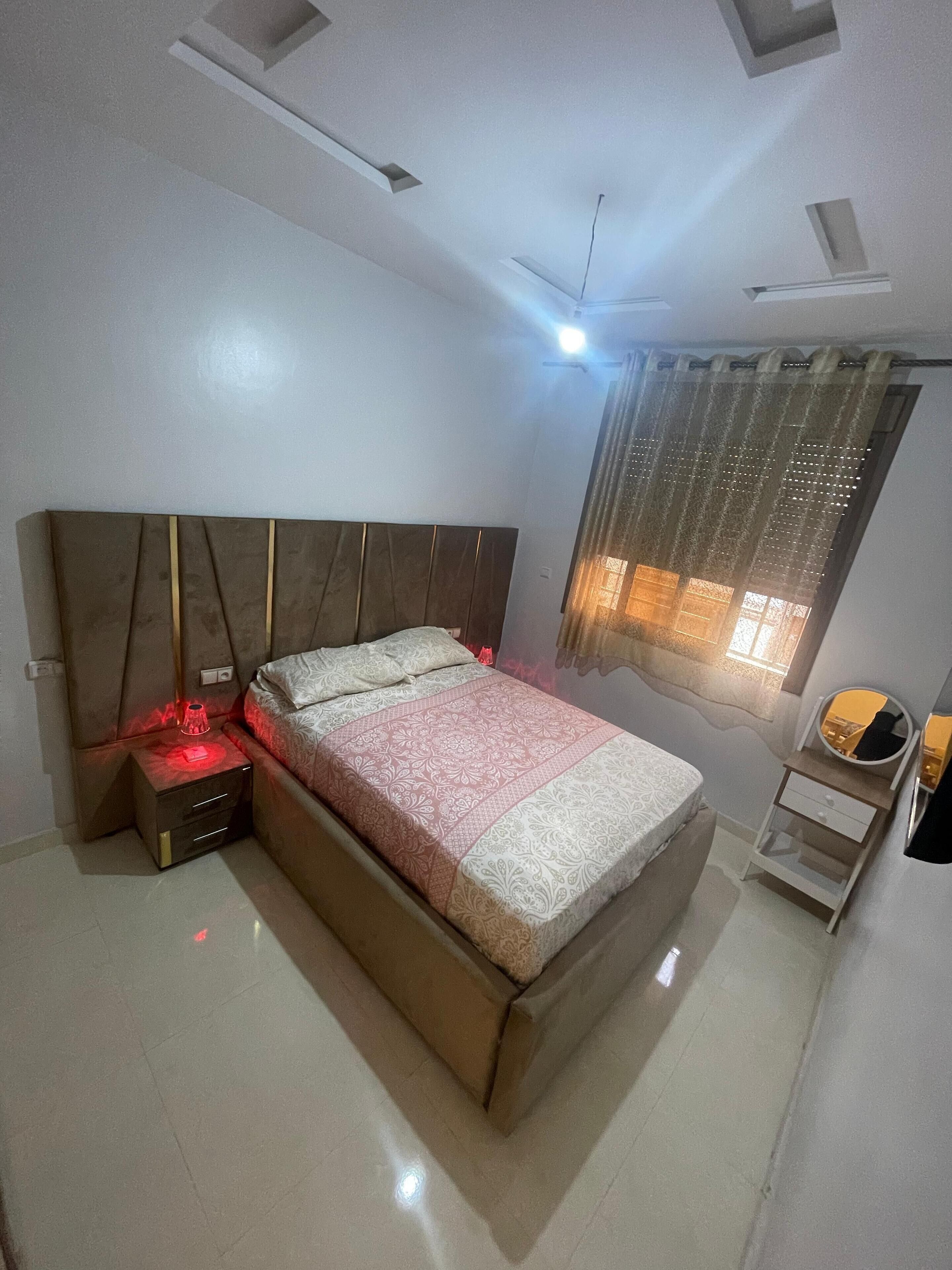 A bedroom with a bed, bedside table, nightstand, ceiling fan, window with curtains, and a small mirror.