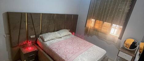 A bedroom with a bed, bedside table, nightstand, ceiling fan, window with curtains, and a small mirror.