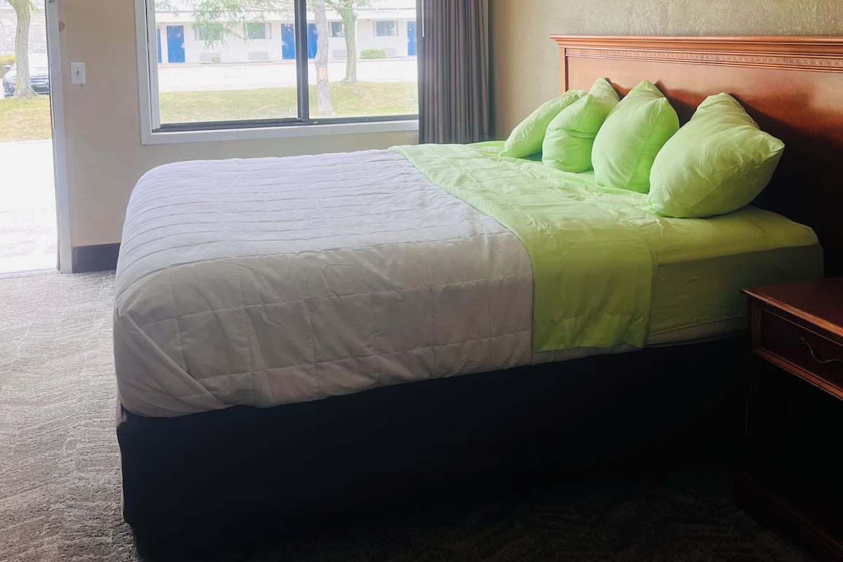 A hotel room with a bed, a nightstand, a ceiling light, a window with curtains, and a view of the outside.