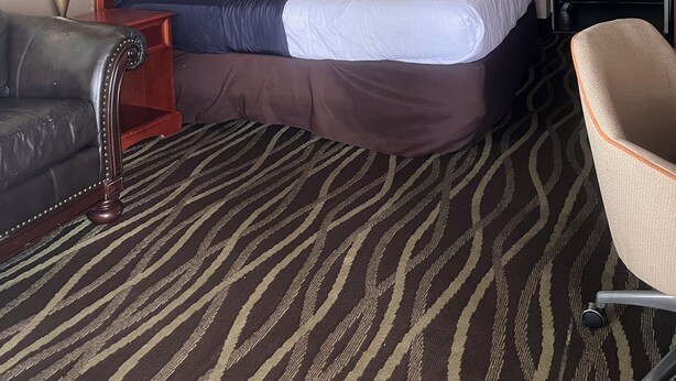 A hotel room with a bed, two bedside lamps, a framed picture on the wall, a patterned carpet, a leather armchair, and a chair. - Stay-Well Inn  (Monroe)