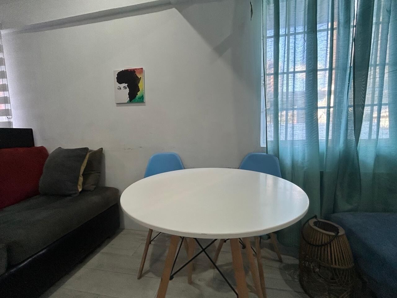 Ein Wohnzimmer mit einem weißen runden Tisch, blauen Stühlen, einem grauen Sofa mit roten und braunen Kissen und einem Gemälde an der Wand.