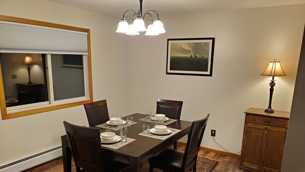 A dining room with a wooden table set for four, chairs, a chandelier, a painting on the wall, and a lamp on a side table. - Tomar Park Apt 8 - KP101APT8 (Sioux Falls)
