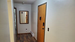 A narrow hallway with wooden flooring, a wooden door with a keyhole, and a wall-mounted mirror. - Tomar Park Apt 8 - KP101APT8 (Sioux Falls)