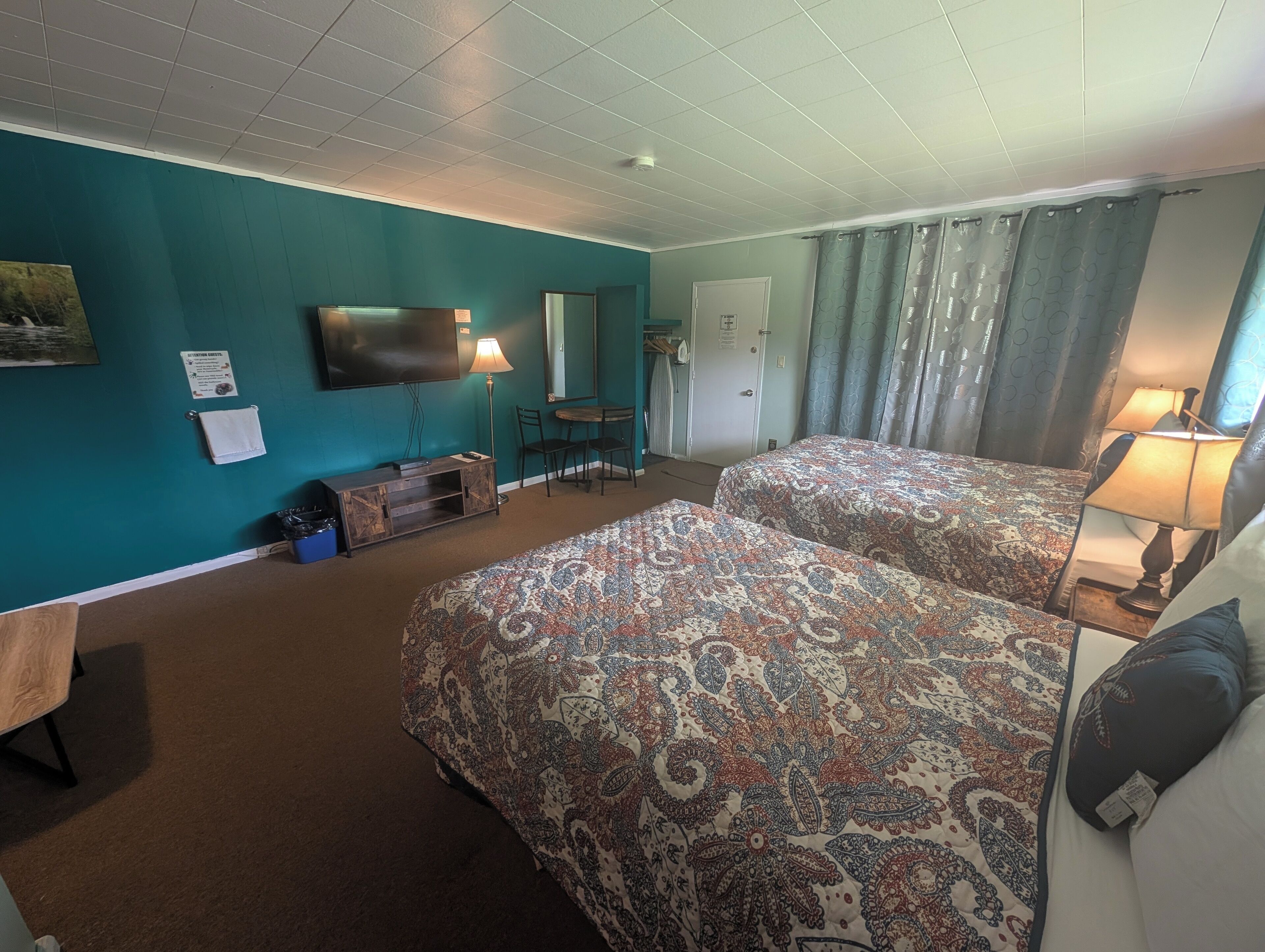 Habitación de hotel con paredes color turquesa, dos camas con cubrecamas estampados, un televisor de pantalla plana, un escritorio con lámpara, una silla y una pequeña mesa.