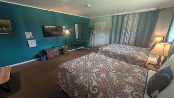 A hotel room with teal walls, two beds with patterned bedspreads, a flat-screen TV, a desk with a lamp, a chair, and a small table.