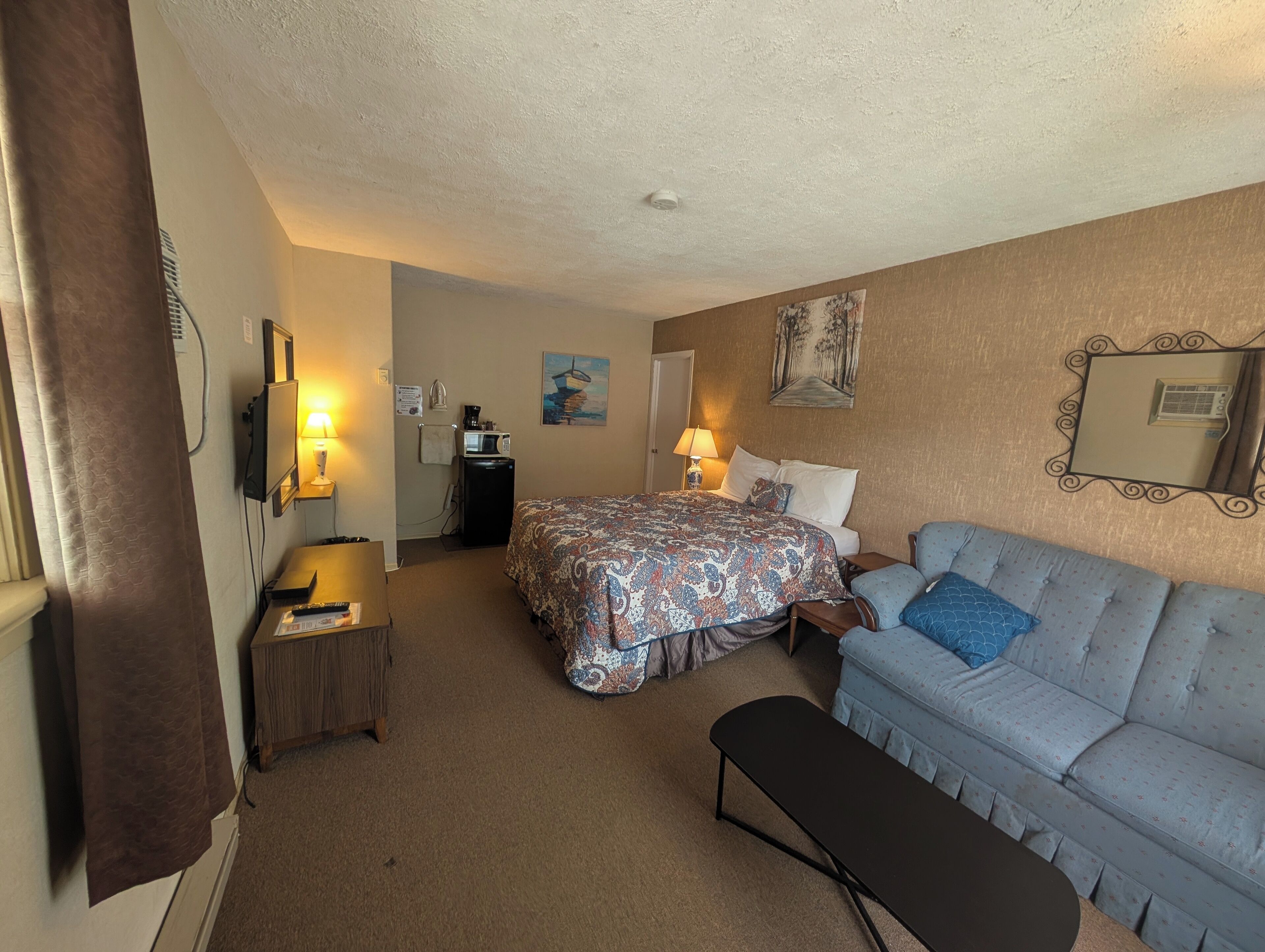 A hotel room with a bed, a sofa, a desk with a lamp, a small kitchenette, and a mirror on the wall.