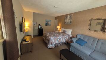 A hotel room with a bed, a sofa, a desk with a lamp, a small kitchenette, and a mirror on the wall.