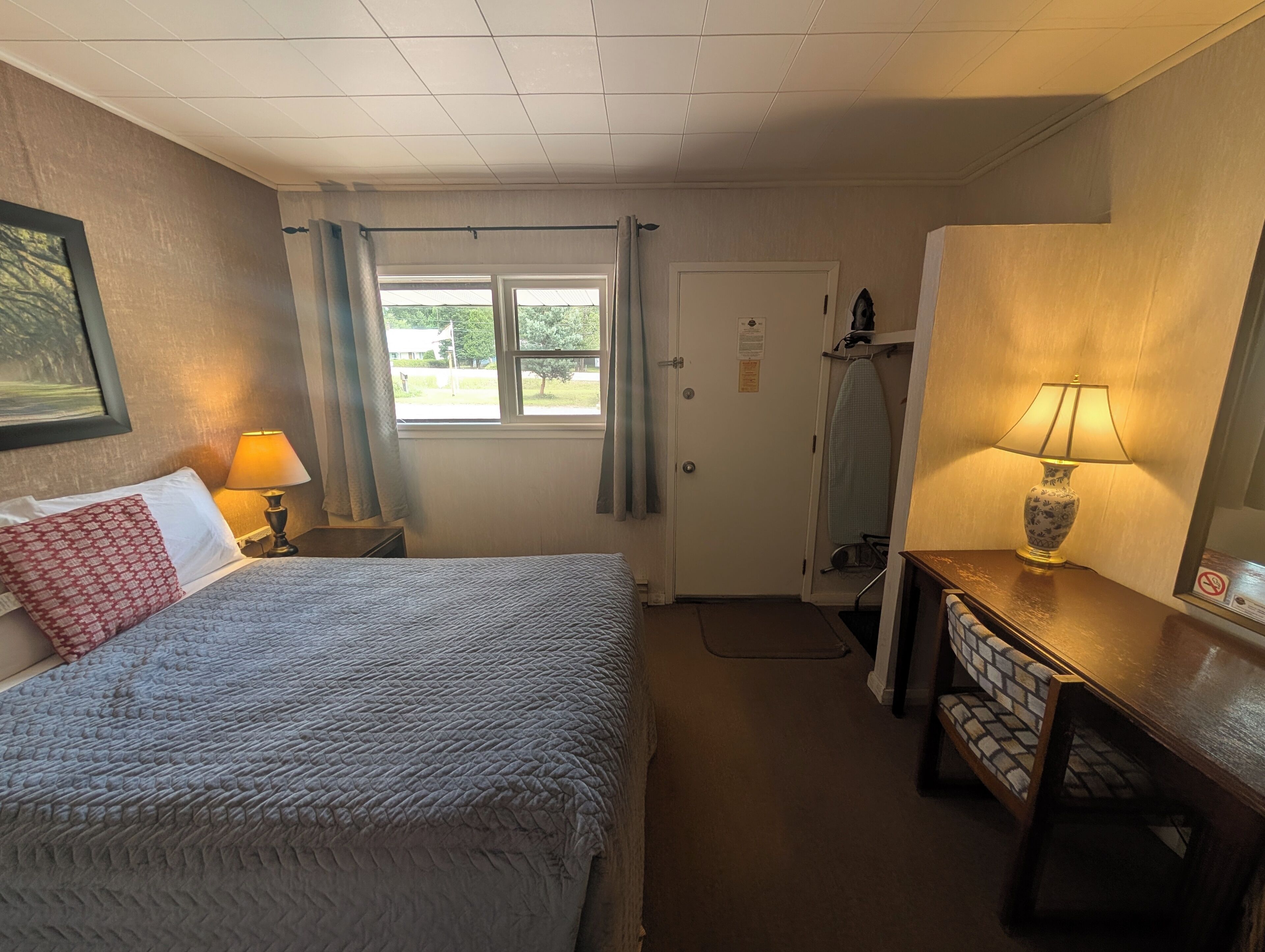 Une chambre d’hôtel avec un lit, un bureau, une lampe, une fenêtre et une porte.