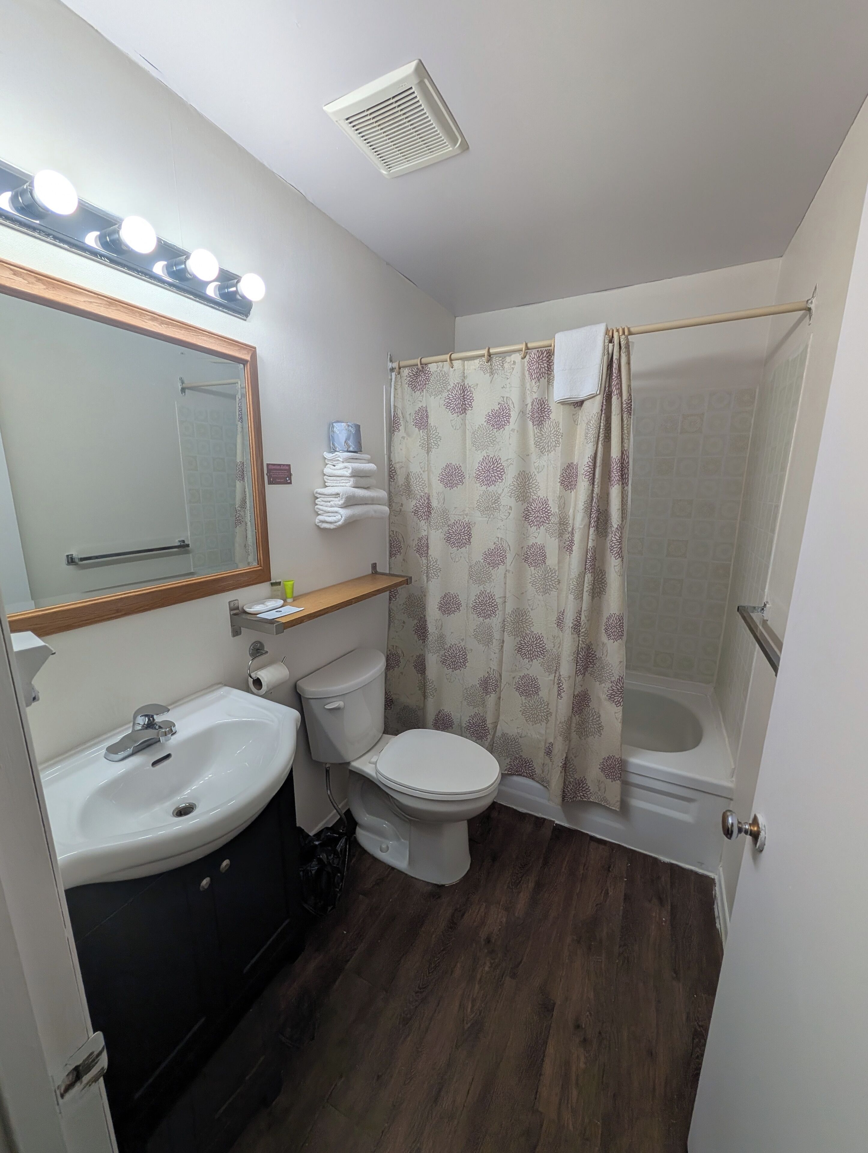 Une salle de bain équipée d’un lavabo, de toilettes et d’une douche. Un miroir avec des lumières est placé au-dessus du lavabo, et un porte-serviettes contient des serviettes pliées.
