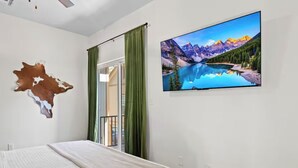 Une chambre à coucher avec un lit, un téléviseur fixé au mur qui diffuse une image d’un lac de montagne, une décoration murale en peau de vache et une porte coulissante avec des rideaux verts.