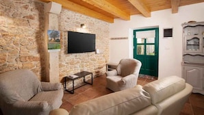 Un salón acogedor con paredes de piedra, vigas de madera en el techo y una puerta verde. Hay dos sillones color beige y un sofá de cuero.