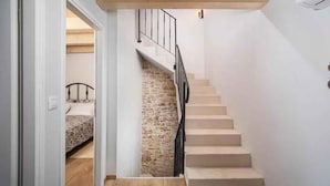 Una escalera con un muro de piedra a un lado y piso de madera al otro, que conduce a un dormitorio con cama y mesita de noche.
