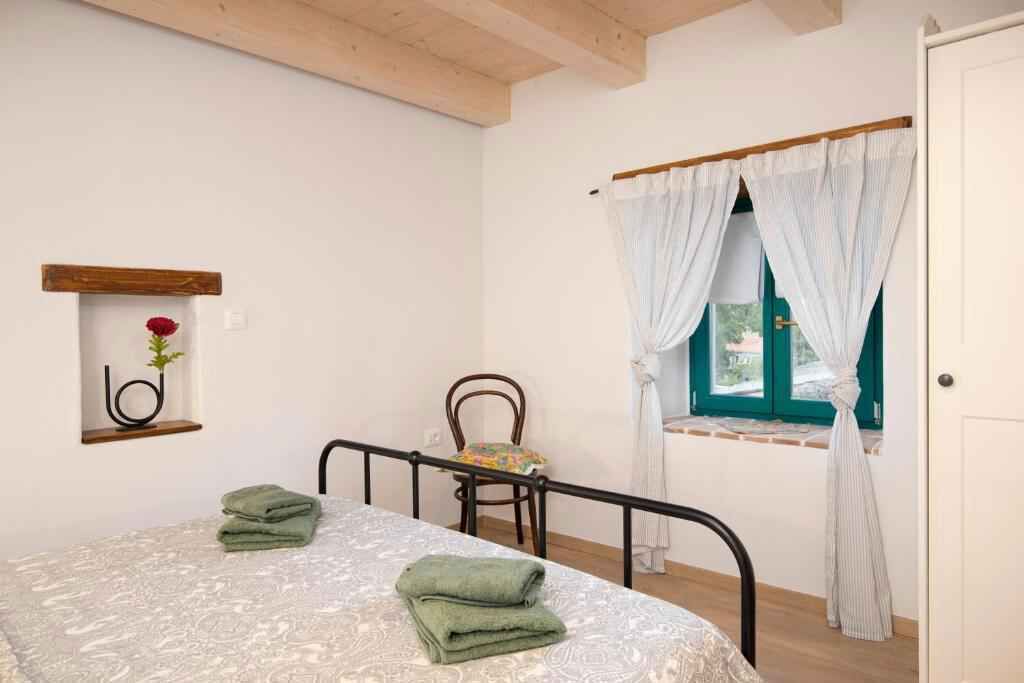 Una camera da letto con un letto, una sedia, una finestra con tende, uno scaffale di legno con un fiore e una porta.