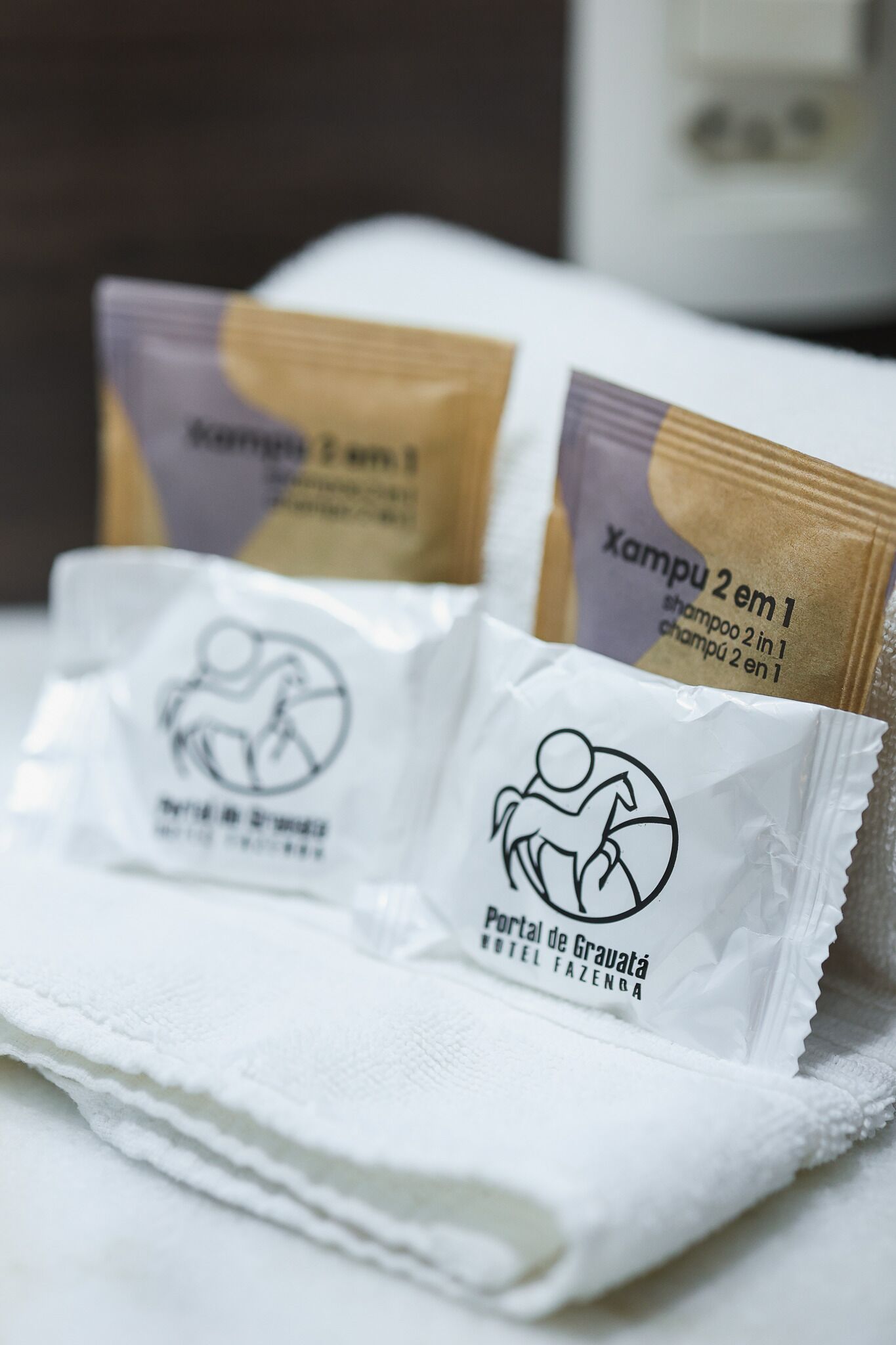 Two shampoo sachets and a towel with a logo on a hotel bed.