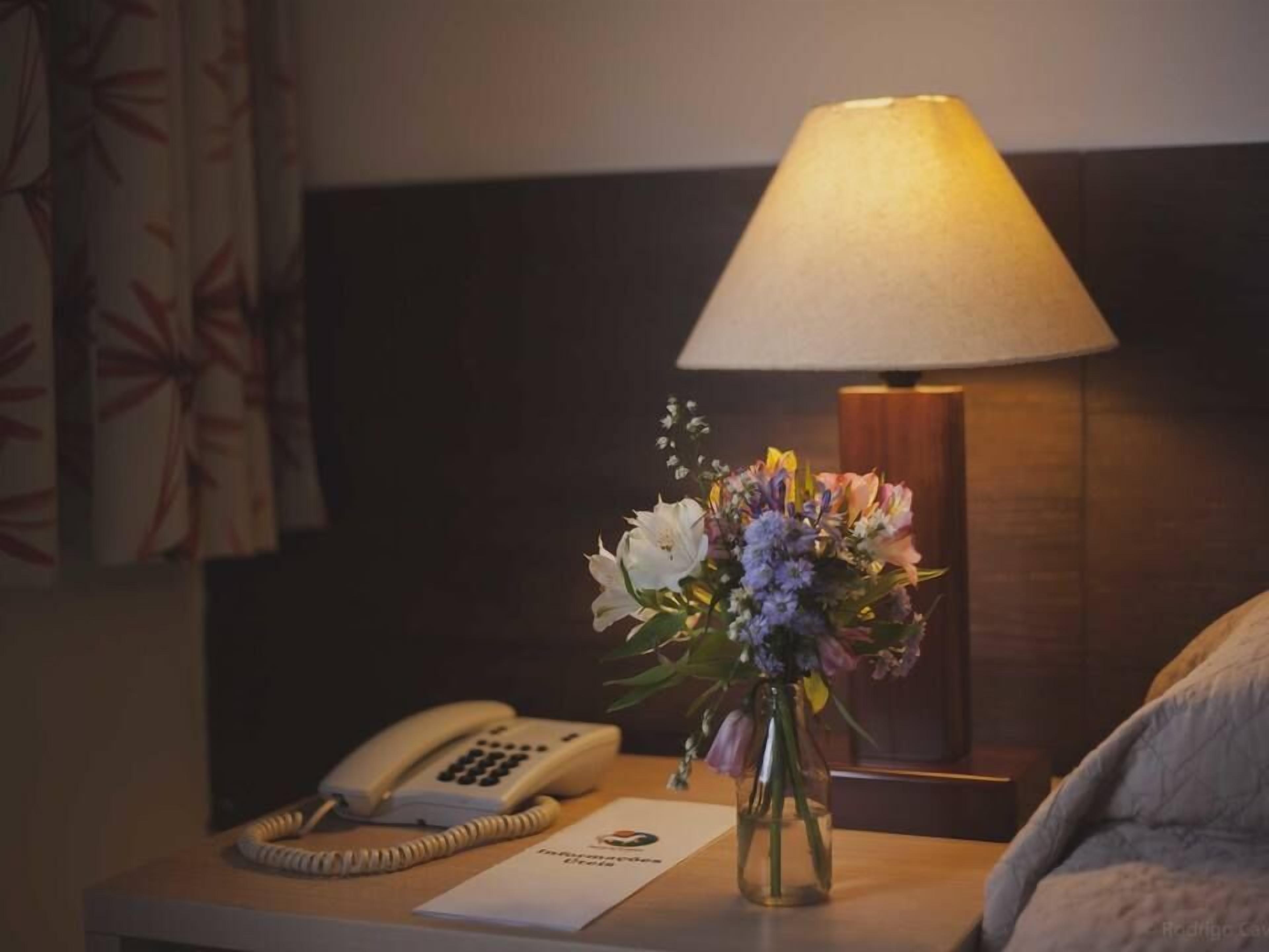 Habitación de hotel con una mesita de noche que incluye una lámpara, un teléfono y un arreglo floral.