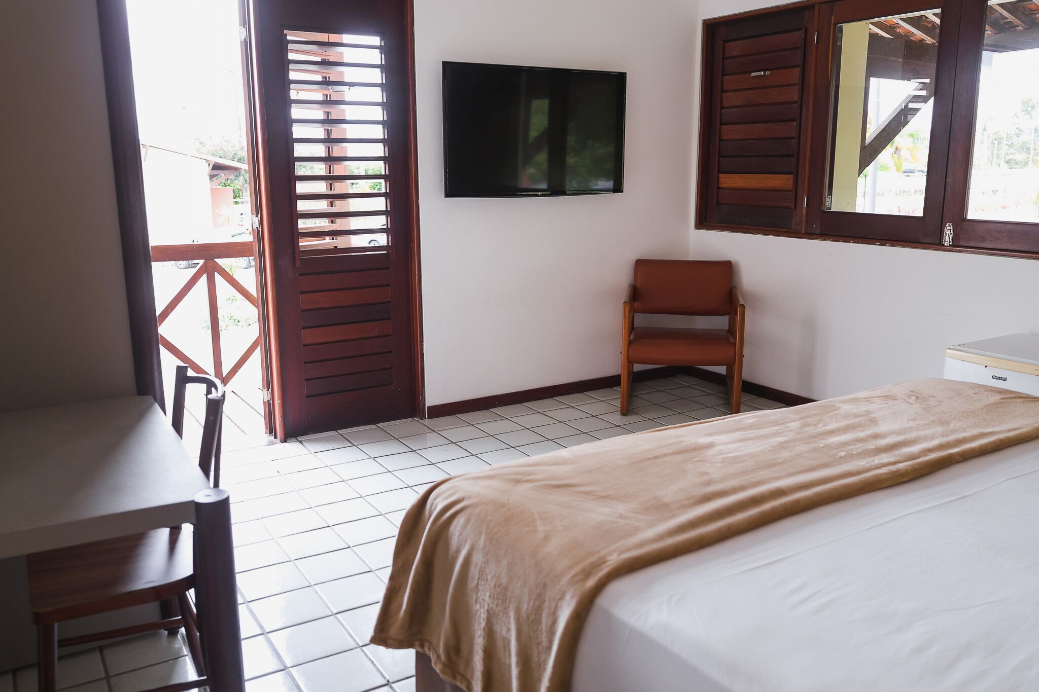 Quarto com cama, escrivaninha, cadeira, televisão e janela com persianas de madeira.