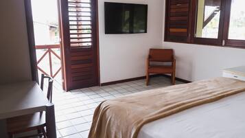Quarto com cama, escrivaninha, cadeira, televisão e janela com persianas de madeira.