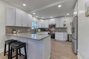 A modern kitchen with white cabinets, a large island with stools, stainless steel appliances, and a window providing natural light.