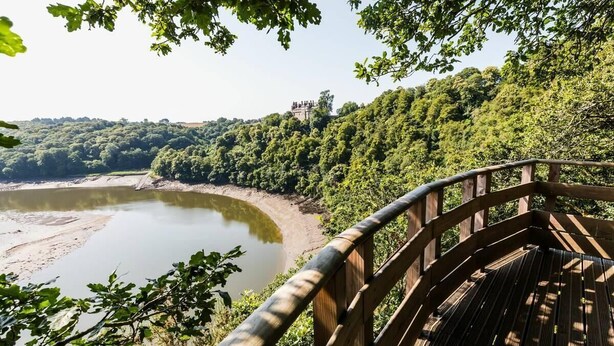 Una terraza de madera con vistas a un río, una ladera boscosa y una estructura que parece un castillo a lo lejos.
