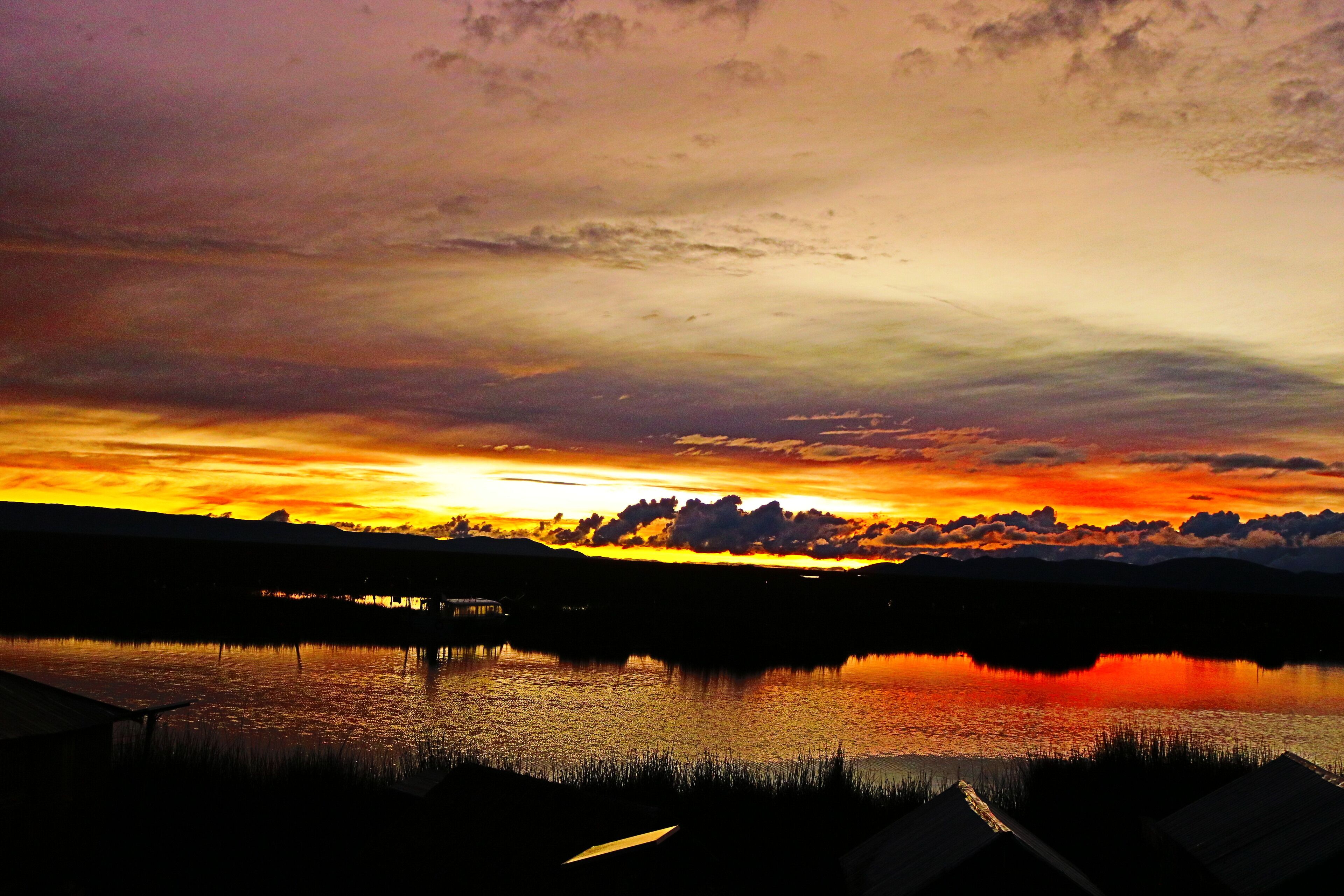Un atardecer sobre un lago tranquilo con un muelle y montañas distantes.