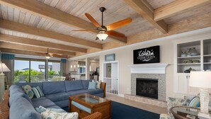 A spacious living room with a large sofa, a coffee table, and a fireplace. There is a ceiling fan, a sign reading "Coastal Carolina," and a view of the outdoors. - Point of View: Saltwater Pool, Hot Tub, Amazing Ocean and Sound Views (Duck)
