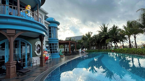 Una amplia piscina curva con piso de baldosas, rodeada de palmeras y un edificio de arquitectura azul y marrón.