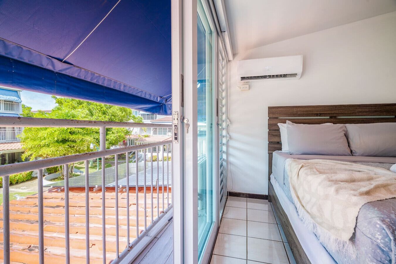 Ein Balkon mit Blick auf die Nachbarschaft, ein Bett mit Holz Kopfteil und eine Klimaanlage an der Wand.