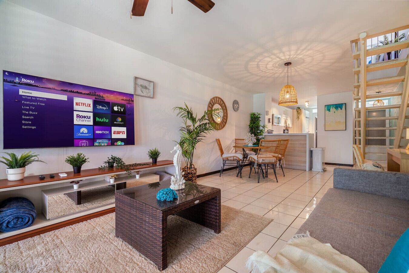 A modern living room with a flat-screen TV displaying a Roku interface, a dining area with wicker chairs, and a kitchen visible in the background.