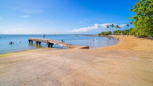 Una playa con un muelle de hormigón que se adentra en el agua, palmeras y un cielo azul despejado.