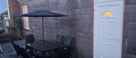 An outdoor patio with a table and chairs, a black umbrella, and a white door with a yellow window.